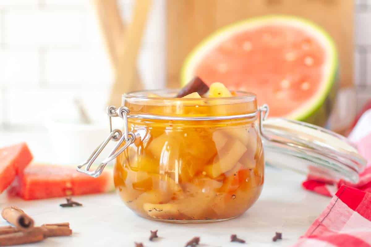 A glass jar filled with pickled fruit pieces and spices sits on a table, with a sliced watermelon and various spices in the background.