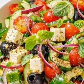 A bowl of Greek salad with cherry tomatoes, cucumber, feta cheese, black olives, red onion, green bell pepper, and fresh mint leaves, seasoned with herbs.