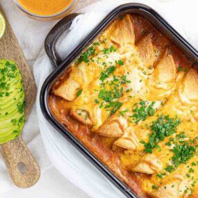 A baking dish of cheesy chicken enchiladas topped with chopped herbs, next to sliced avocado, lime wedges, and a bowl of sauce on a wooden board—chicken meals like this keep my weeknight dinners satisfying and simple.