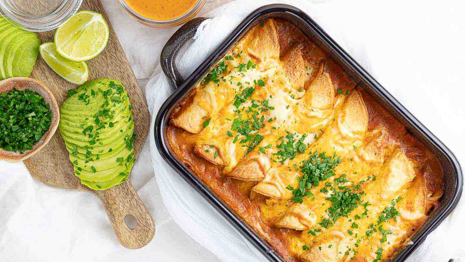 A baking dish of cheesy chicken enchiladas topped with chopped herbs, next to sliced avocado, lime wedges, and a bowl of sauce on a wooden board—chicken meals like this keep my weeknight dinners satisfying and simple.