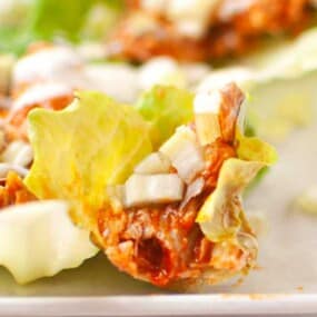Close-up of a lettuce leaf topped with shredded meat in sauce, diced onions, and a drizzle of white dressing, on a white plate.
