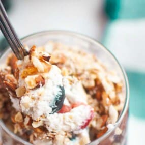A spoonful of dessert with whipped cream, chopped nuts, and fruit held above a glass, with a green checkered cloth in the background.