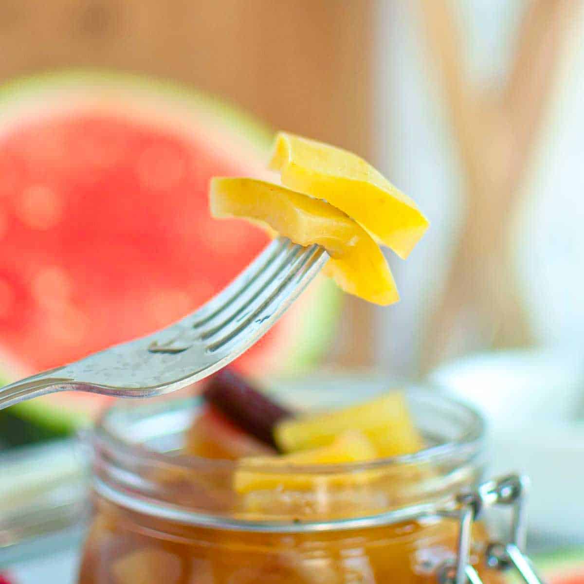 A fork holds slices of yellow pickled watermelon rind above an open glass jar, with a blurred watermelon in the background.