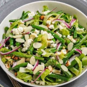 A bowl of mixed green salad with green beans, sliced red onions, almonds, crumbled cheese, and lettuce, served on a gray plate with a fork.