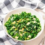 A white bowl filled with chopped kale salad topped with sliced almonds and grated cheese, placed on a plate next to a checkered napkin.