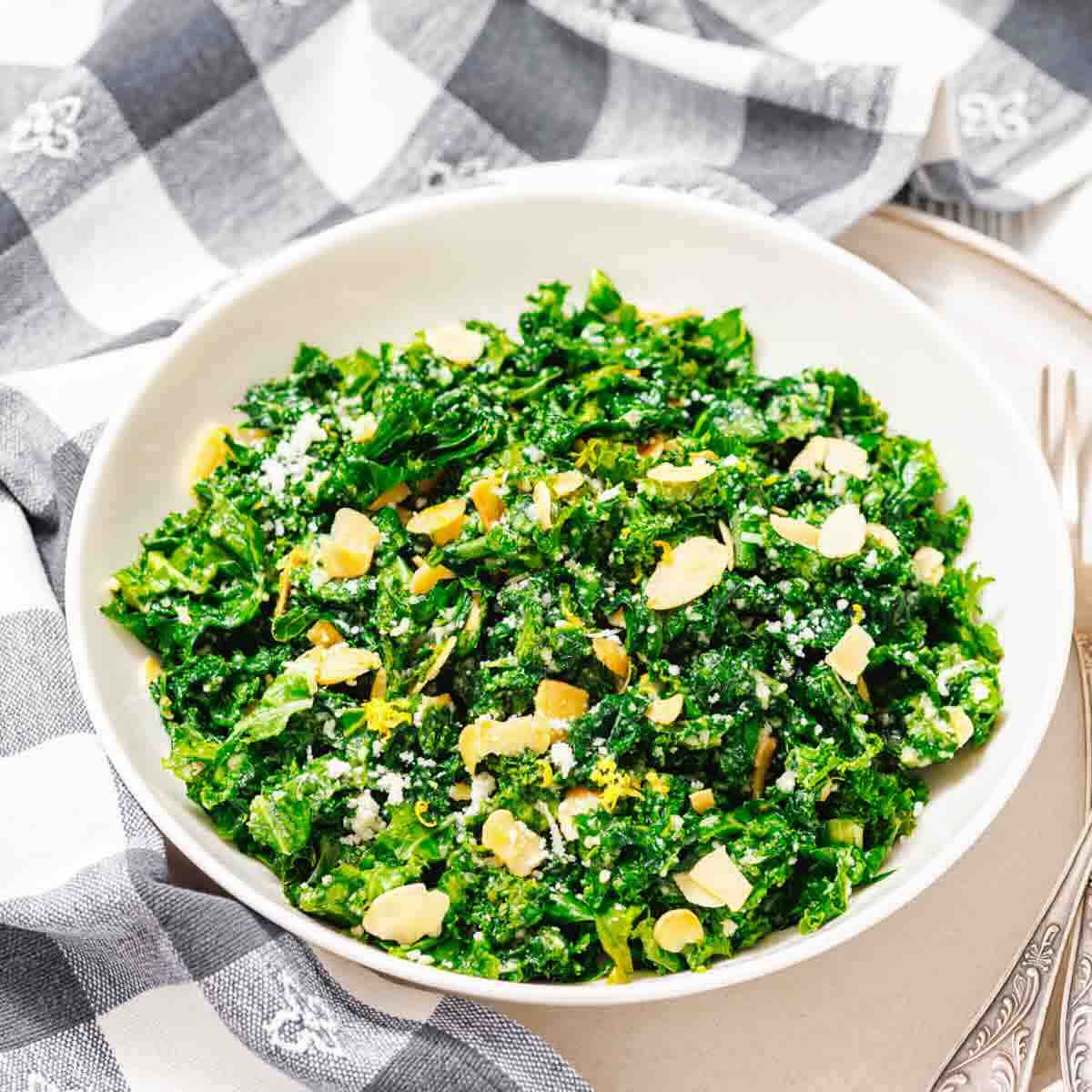 A white bowl filled with chopped kale salad topped with sliced almonds and grated cheese, placed on a plate next to a checkered napkin.