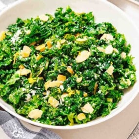 A white bowl filled with chopped kale salad topped with sliced almonds and grated cheese, placed on a plate beside two forks and a checkered napkin.