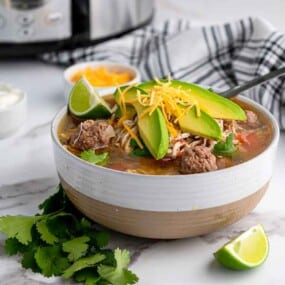 A bowl of beef soup topped with sliced avocado, shredded cheese, and cilantro sits on a marble counter, with lime wedges, a spoon, and a slow cooker in the background.