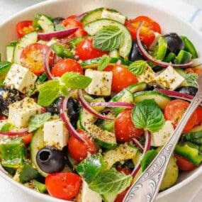 A bowl of Greek salad with cherry tomatoes, cucumber, olives, red onion, feta cheese, green pepper, and fresh mint leaves, with a fork resting on the side.