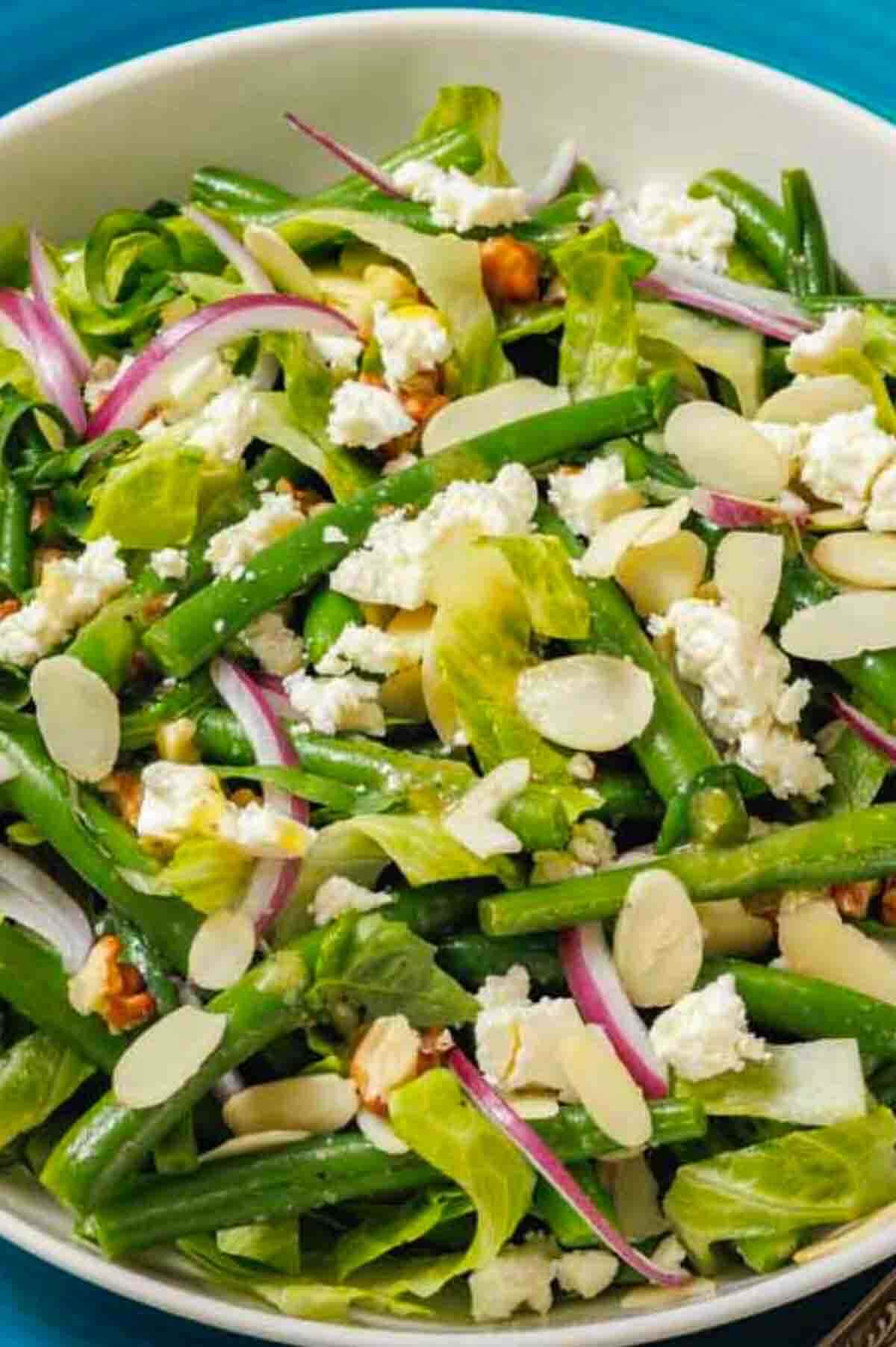 A bowl of salad with green beans, lettuce, red onion slices, feta cheese, almond slices, and walnuts on a blue surface.