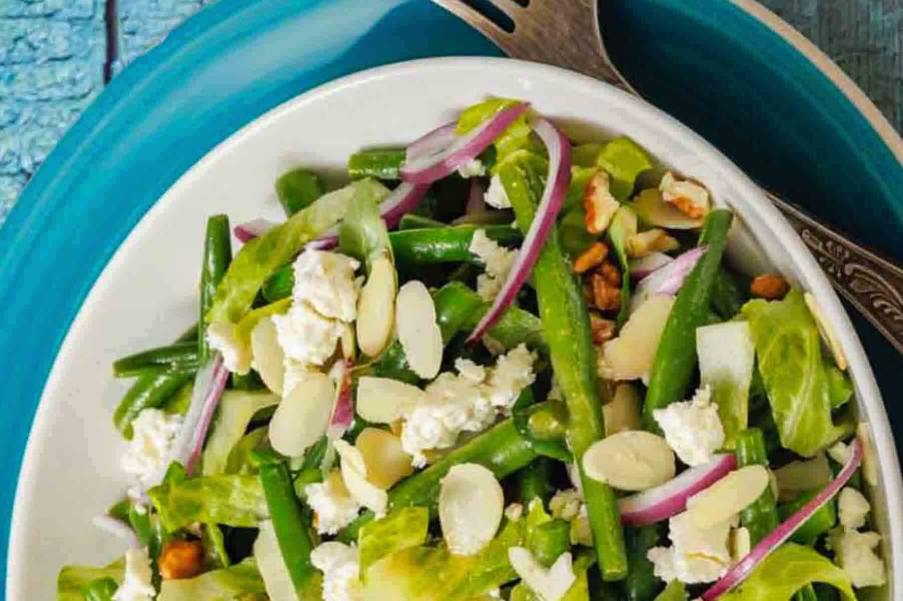 A bowl of walnut goat cheese salad with sliced almonds, red onions, lettuce, walnuts, and crumbled cheese on a blue plate with a fork.