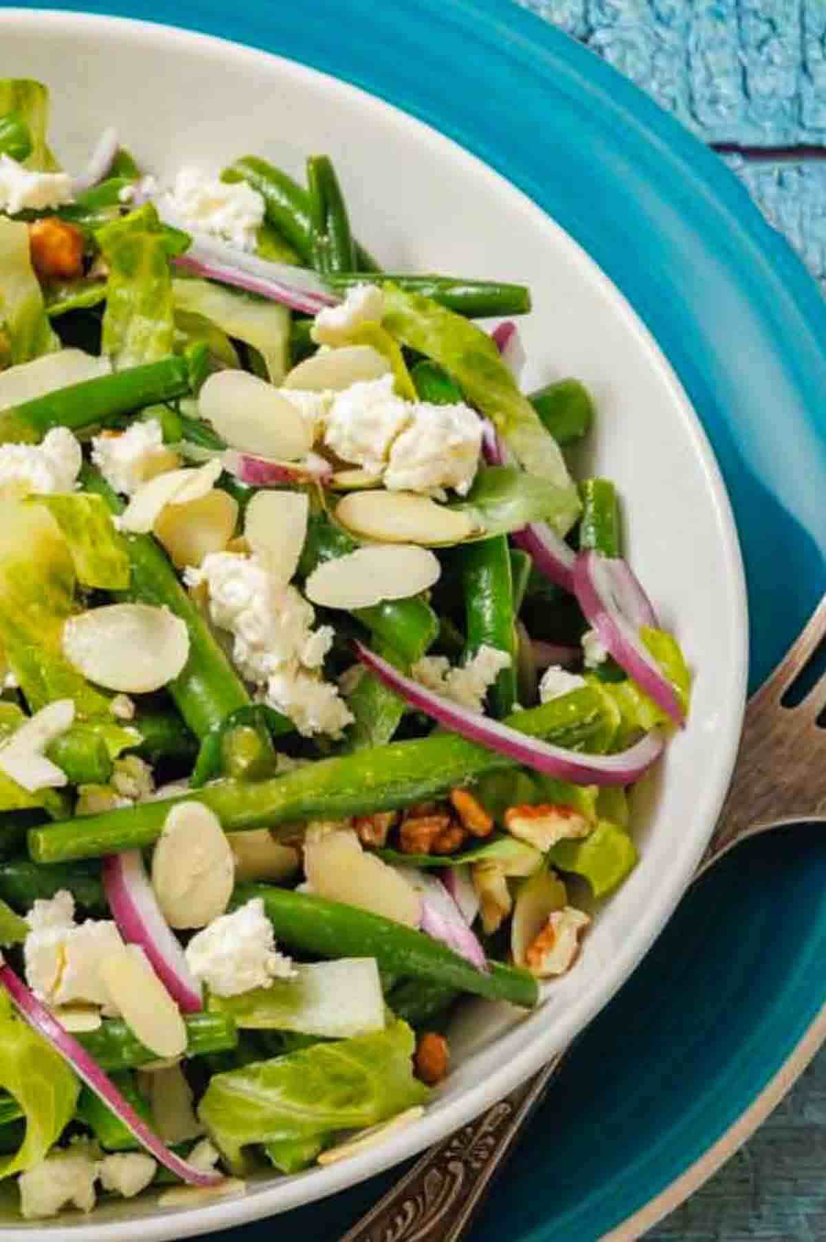 A bowl of green bean salad with sliced almonds, red onion, lettuce, crumbled cheese, and a blue plate underneath.
