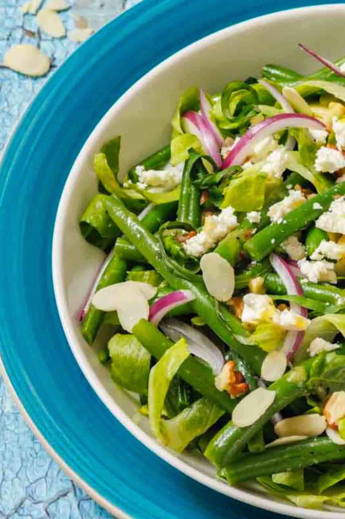 A salad with green beans, lettuce, red onion slices, almond slivers, and crumbled cheese in a white bowl on a blue plate.