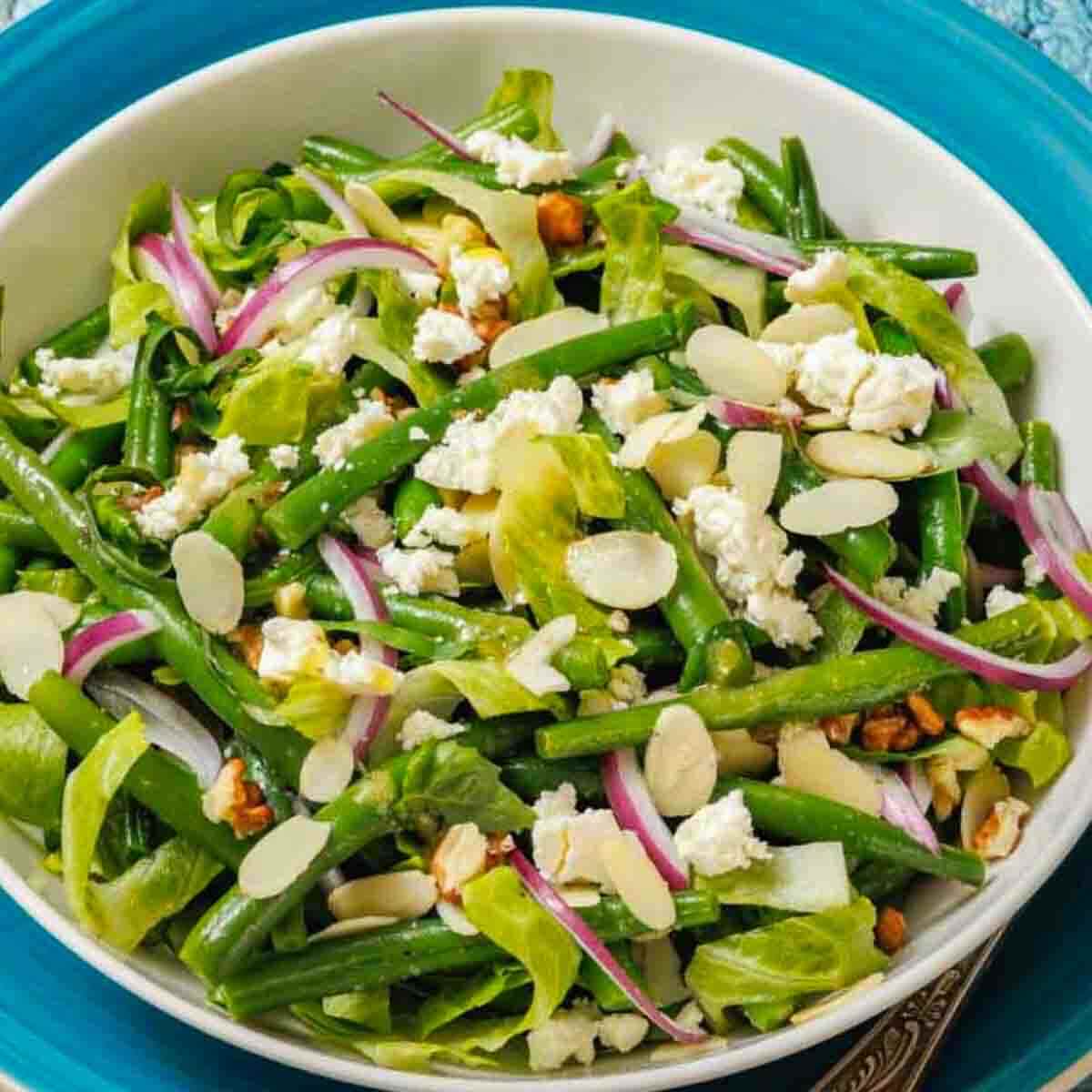 A bowl of green bean salad with lettuce, sliced red onions, feta cheese, almond slices, and walnuts on a blue plate.