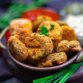 A bowl of breaded and fried mushrooms garnished with parsley, with dipping sauces and fresh herbs in the background.