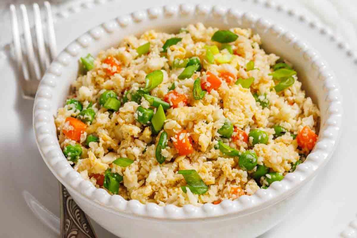 A white bowl filled with cauliflower fried rice mixed with peas, diced carrots, and chopped green onions, placed on a white plate with a fork beside it.