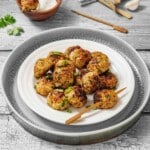A plate of cooked chicken meatballs garnished with herbs, served with wooden skewers on a rustic wooden table. A bowl of dipping sauce and some fresh herbs are in the background.