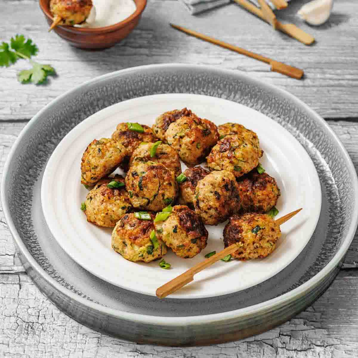A plate of cooked chicken meatballs garnished with herbs, served with wooden skewers on a rustic wooden table. A bowl of dipping sauce and some fresh herbs are in the background.