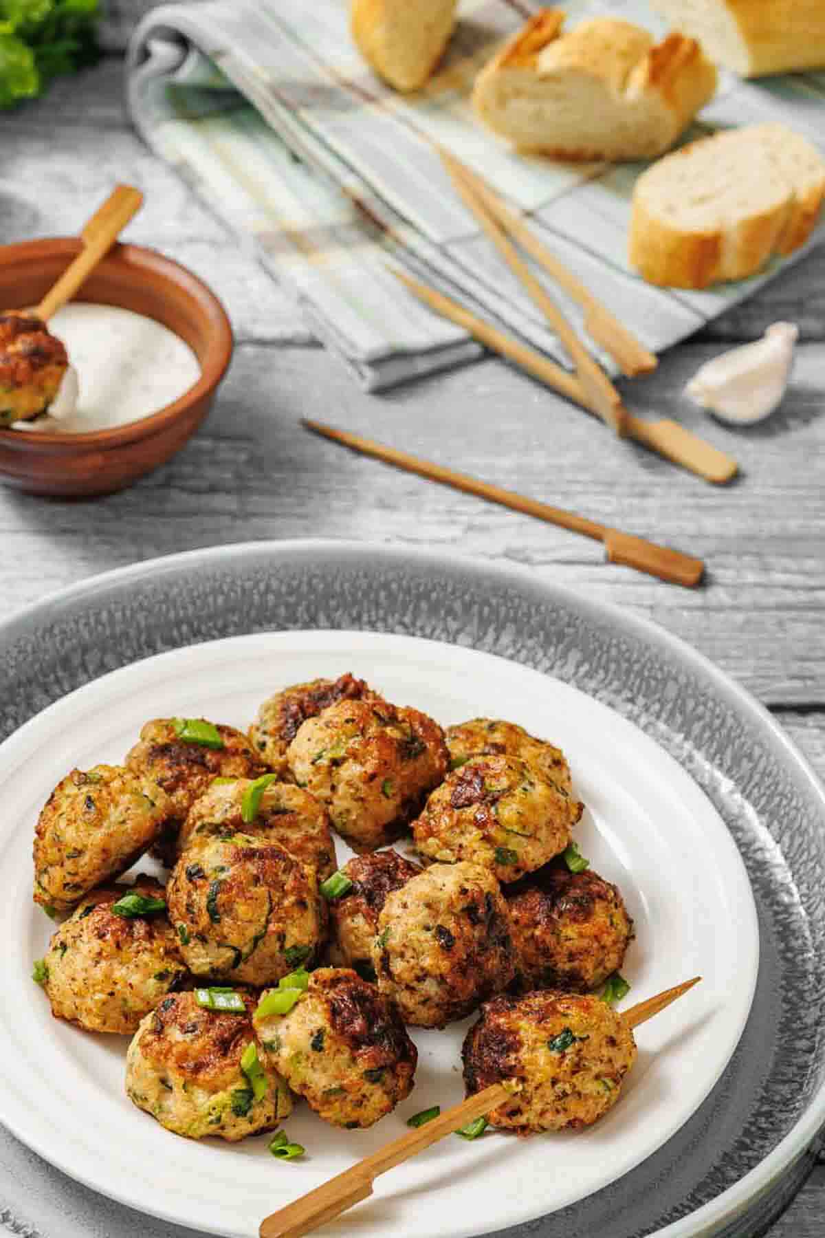 A plate of grilled meatballs garnished with herbs, served with dipping sauce and slices of bread on a wooden table.