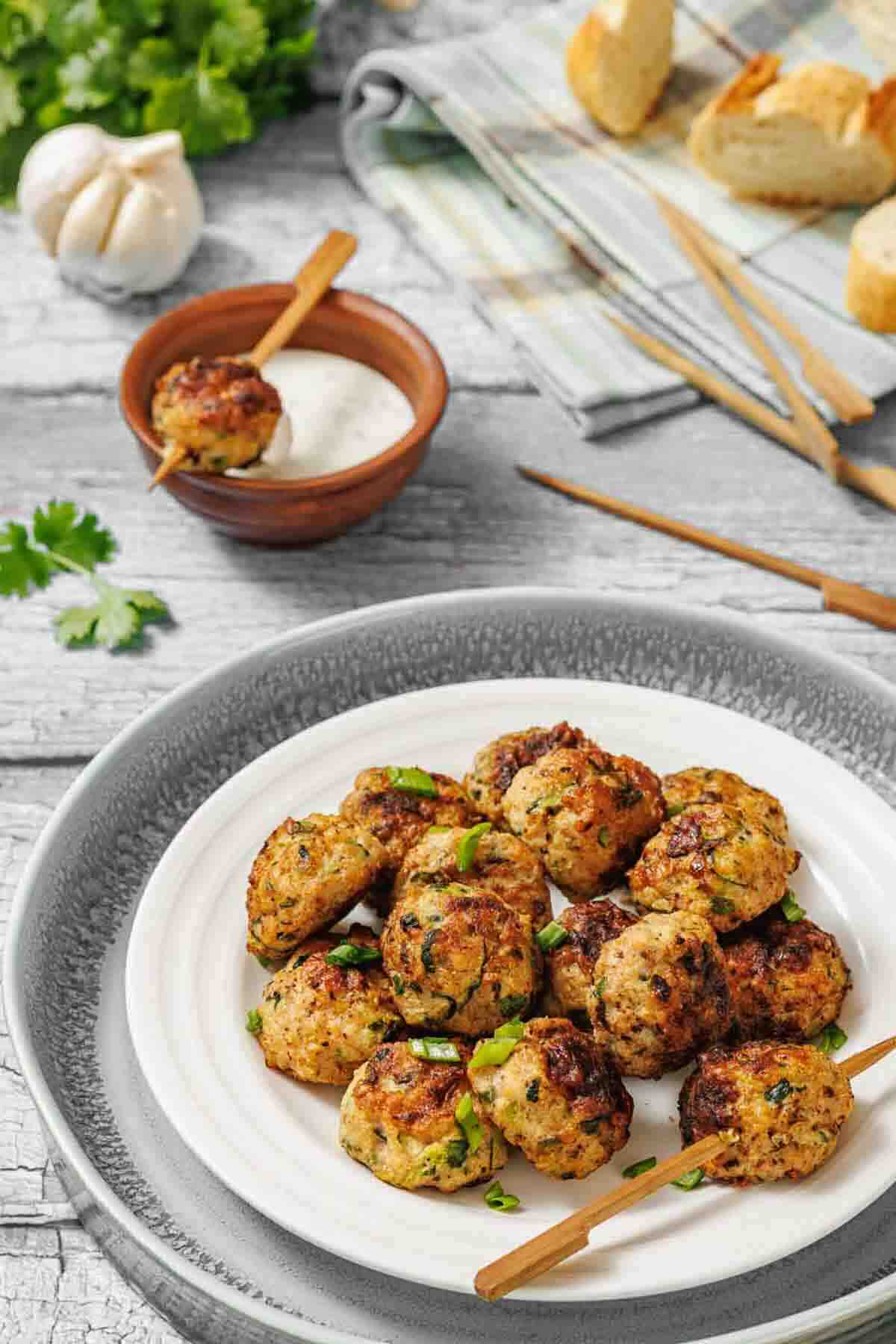 A plate of cooked meatballs garnished with herbs, served with wooden skewers and a bowl of dipping sauce; bread and garlic visible in the background.