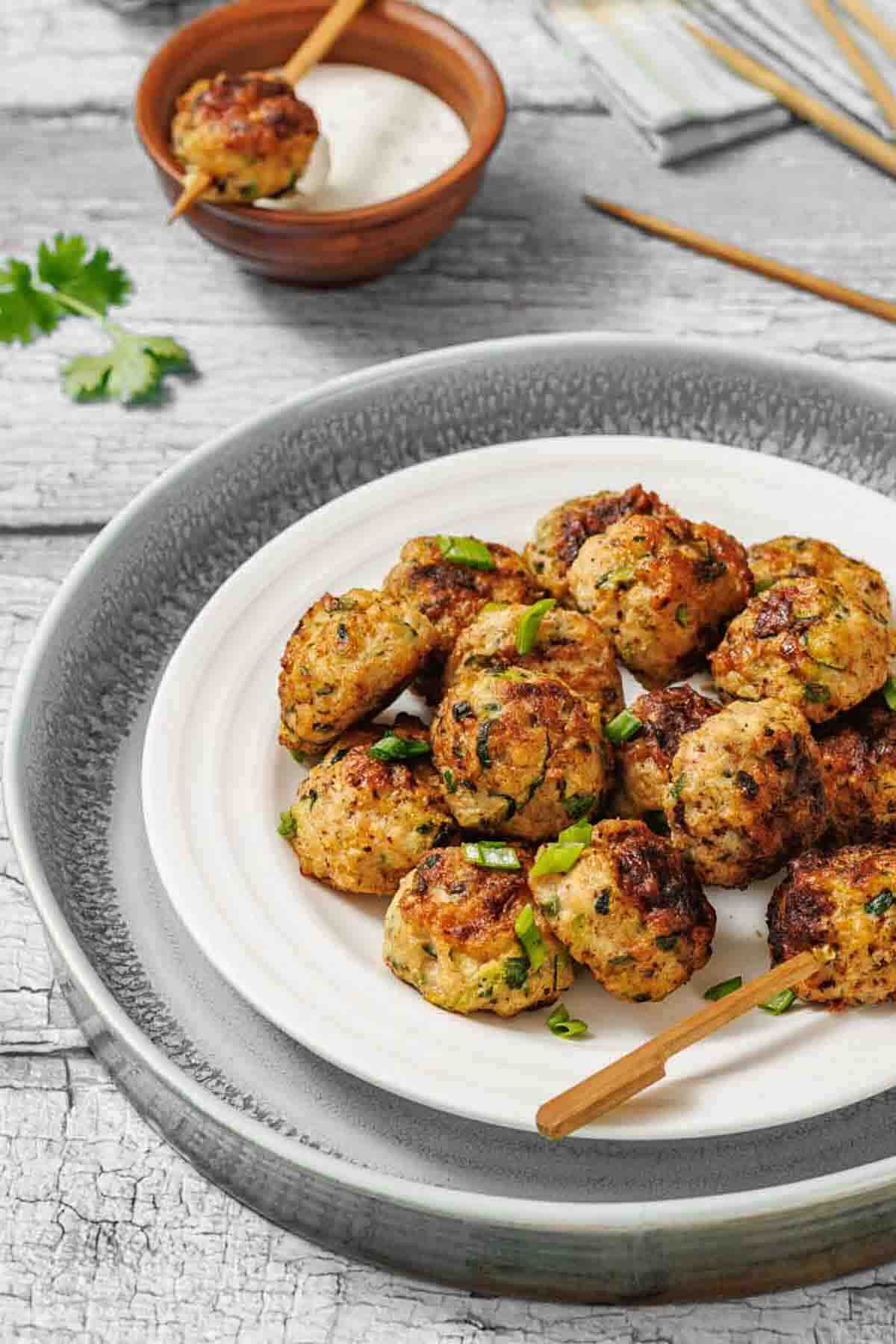 A plate of browned meatballs garnished with chopped herbs, with skewers and a small bowl of dipping sauce in the background.