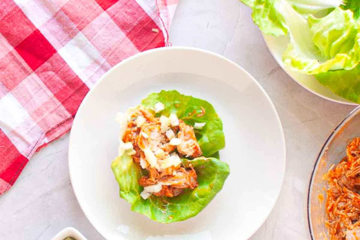 A white plate with a lettuce leaf topped with shredded chicken and diced onions, next to a red and white checkered cloth and fresh lettuce leaves.