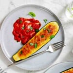 A stuffed and baked zucchini half is served on a gray plate with tomato sauce and garnished with fresh basil, alongside a fork.