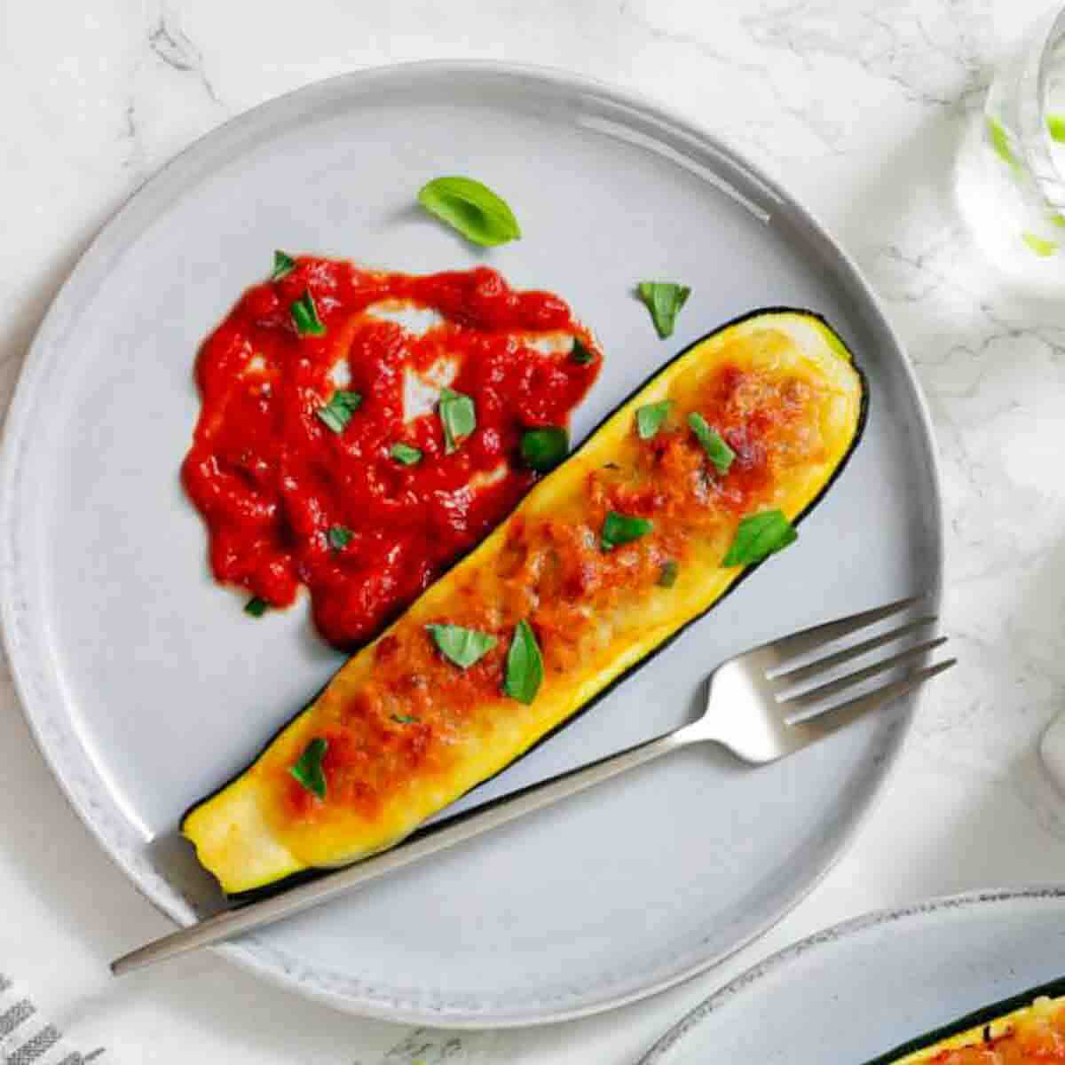 A stuffed and baked zucchini half is served on a gray plate with tomato sauce and garnished with fresh basil, alongside a fork.