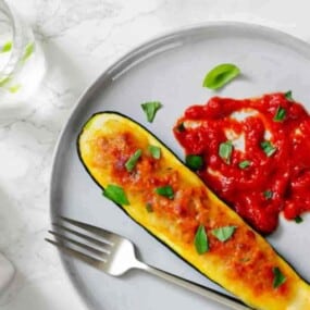 A stuffed zucchini boat topped with melted cheese and herbs, served with a portion of sauce on a gray plate, next to a fork and a glass of water.