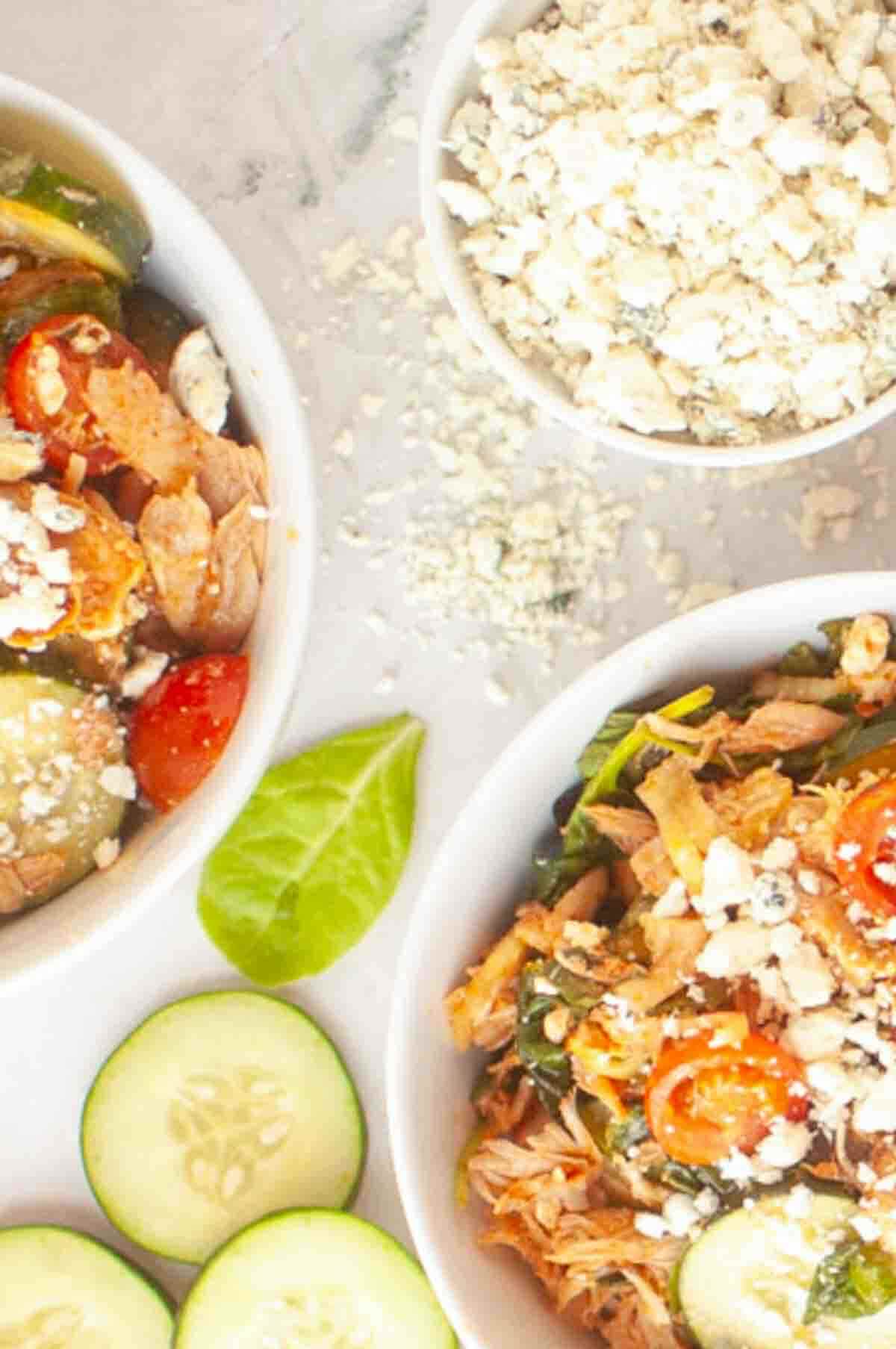 Two bowls of salad with shredded chicken, grape tomatoes, cucumbers, and crumbled cheese, next to a small bowl of more crumbled cheese on a white surface.