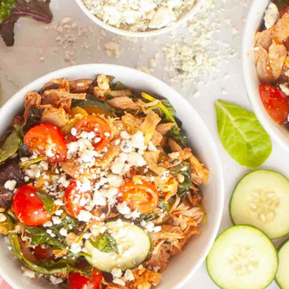 A bowl of salad with shredded meat, cherry tomatoes, spinach, sliced cucumber, and crumbled cheese, with extra cheese and cucumber slices nearby.