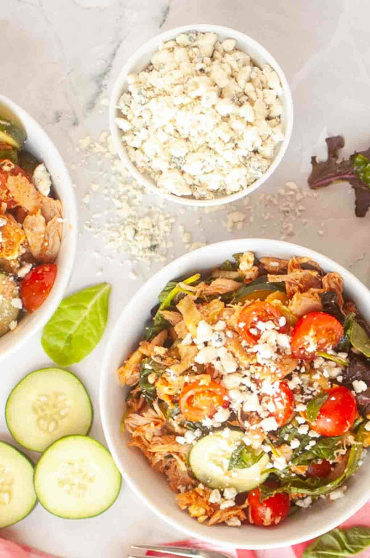 Two bowls of salad with shredded chicken, cherry tomatoes, cucumbers, and crumbled cheese next to a bowl of extra cheese and sliced cucumbers on a marble surface.