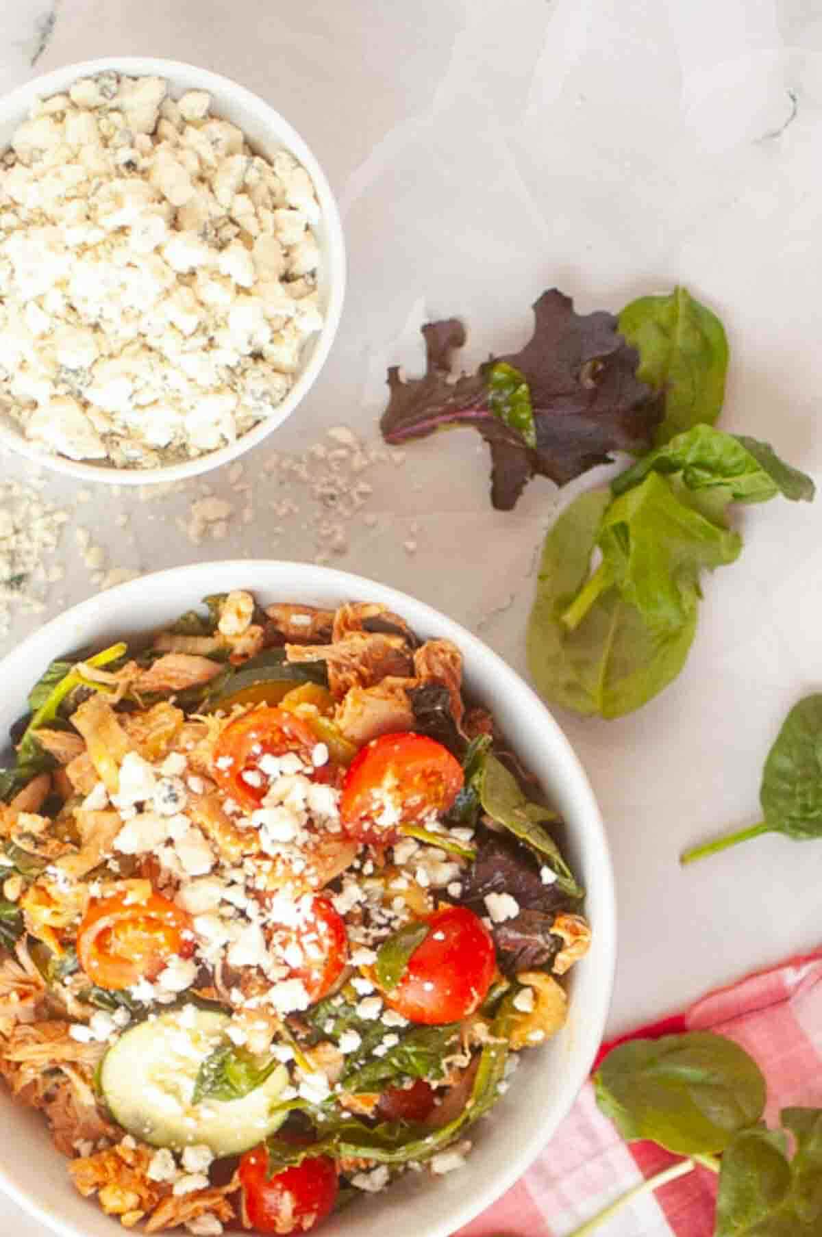 A bowl of salad with cherry tomatoes, cucumbers, shredded meat, greens, and crumbled cheese, next to a small bowl of crumbled cheese and loose salad leaves on a marble surface.