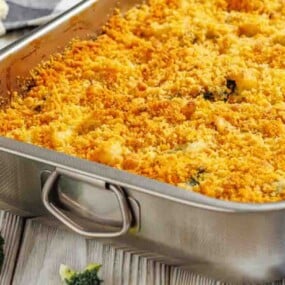 A metal baking dish filled with baked casserole topped with a golden, crispy breadcrumb layer, with broccoli florets visible on the table nearby.