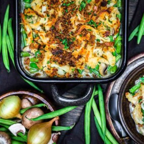 A casserole dish filled with green bean casserole topped with crispy onions, surrounded by fresh green beans, onions, and mushrooms on a dark table.