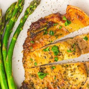 Grilled chicken breast sliced and garnished with herbs, served with spears of asparagus on a white plate, sprinkled with black pepper.