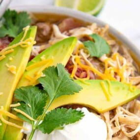 Bowl of shredded chicken soup topped with avocado slices, shredded cheese, sour cream, and cilantro, with a lime wedge in the background.
