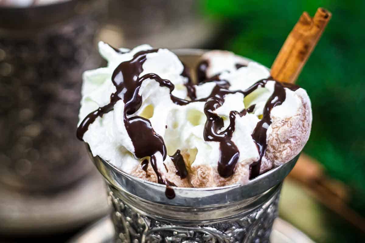 A metal cup filled with whipped cream, hot chocolate, chocolate syrup, and a cinnamon stick garnish.