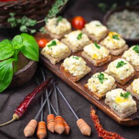 Several pieces of bread topped with a creamy feta and herb eggs, arranged on a wooden board, surrounded by fresh basil, dried chilies, and cherry tomatoes.
