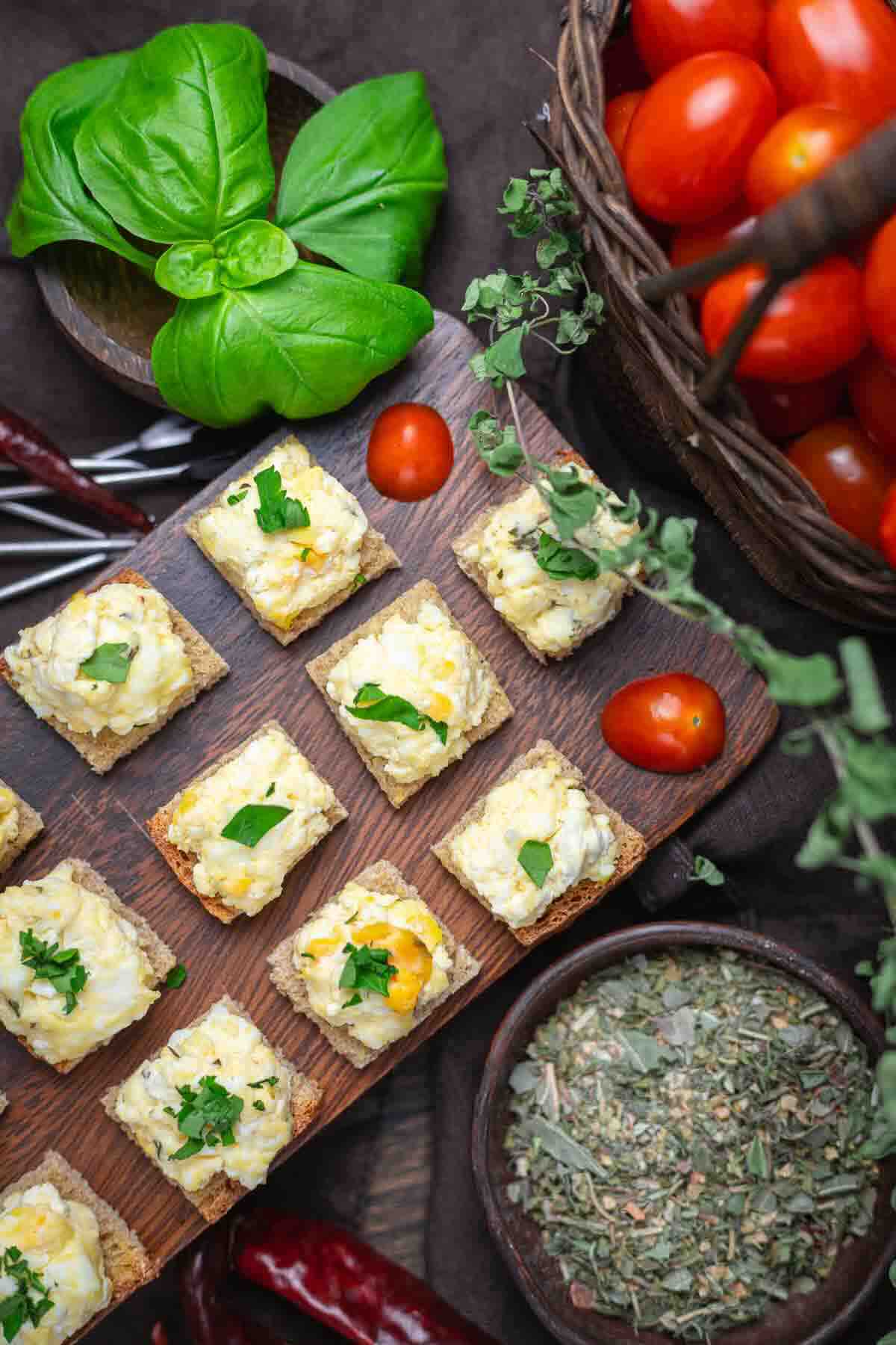 Squares of crackers topped with a creamy spread and herbs are arranged on a wooden board, surrounded by fresh basil, cherry tomatoes, and bowls of dried herbs.