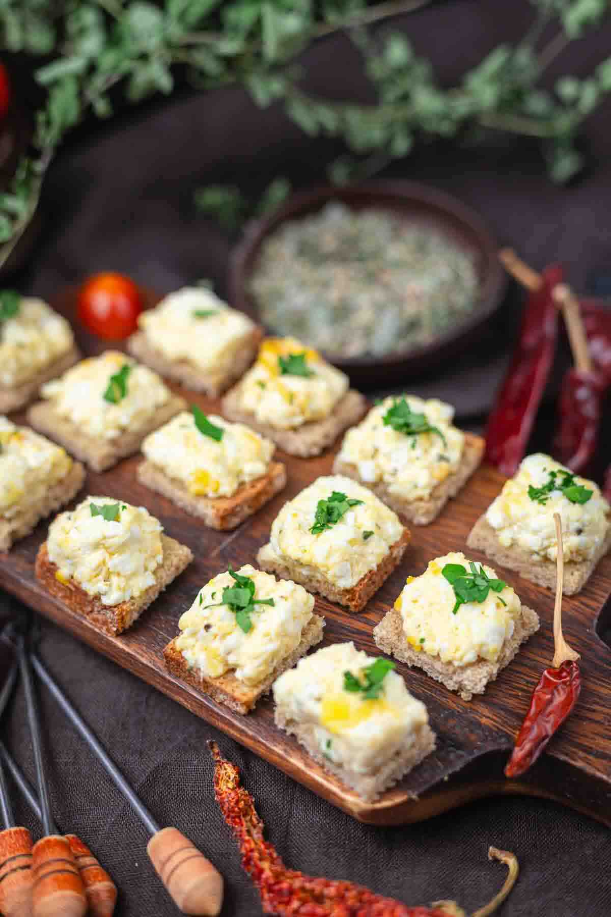 Small pieces of bread topped with an egg salad mixture and garnished with chopped herbs, arranged on a wooden serving board with spices and herbs nearby.