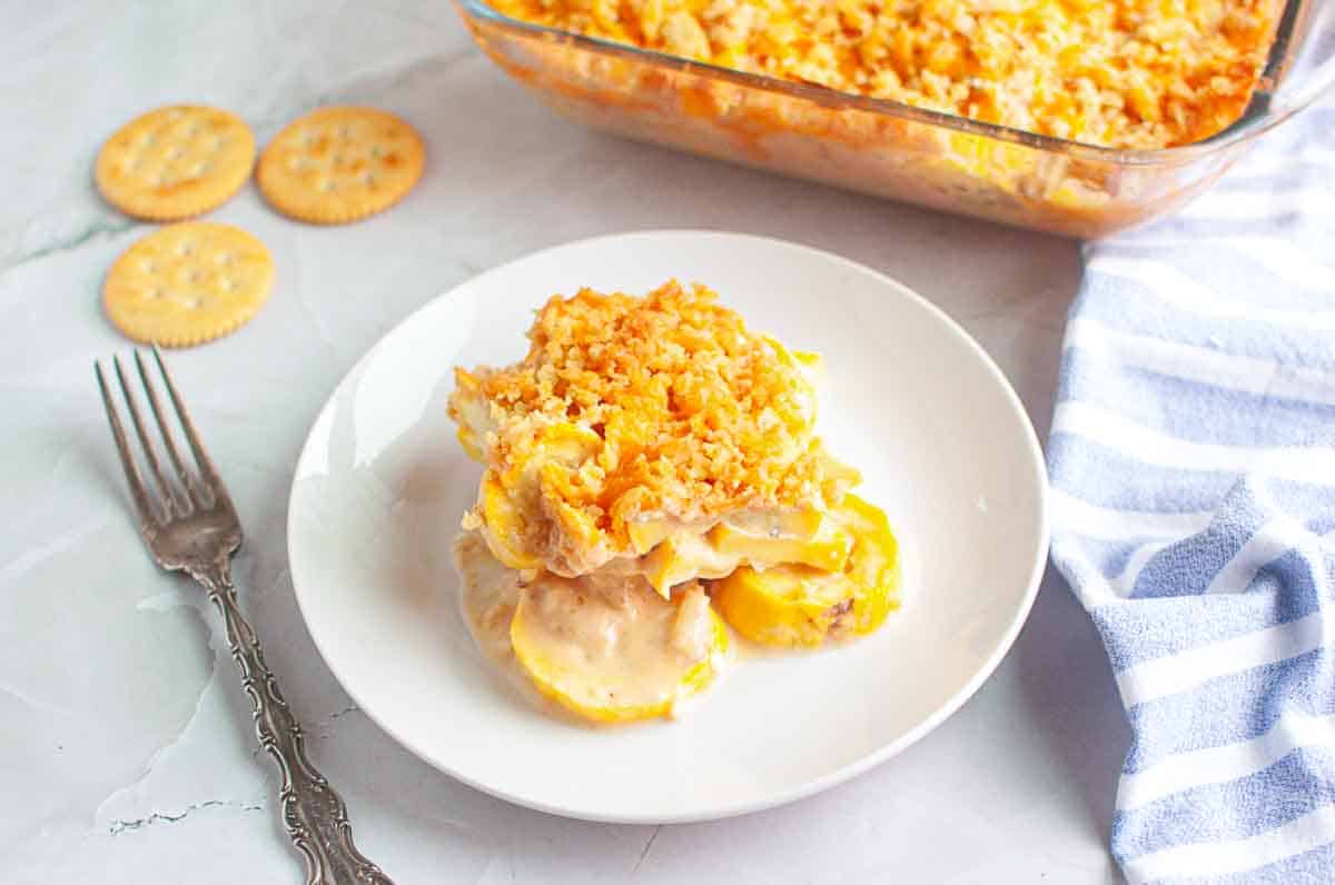 A serving of baked squash casserole with a crumbly topping on a white plate, next to a fork, crackers, and a baking dish on a light surface.