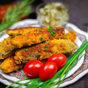 A plate of breaded and fried fish fillets garnished with chives and cherry tomatoes, with herbs, pickles, and a dipping sauce nearby.