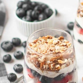A glass filled with layers of yogurt, berries, granola, and chopped nuts, with blueberries and a checkered napkin in the background.