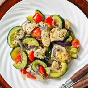 A plate of cooked chicken, zucchini, red bell peppers, and red onions, garnished with herbs, served on a white dish with a brown charger and fork and knife on the side.