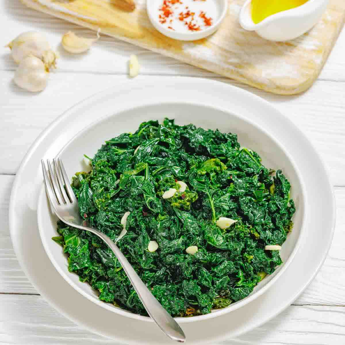 A bowl of cooked chopped spinach garnished with sliced garlic, with a fork on the side; garlic cloves, oil, and spices on a wooden board in the background.