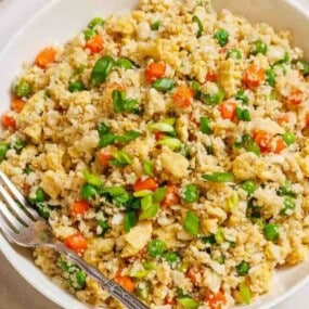 A bowl of cauliflower fried rice with peas, carrots, and chopped green onions, served with a fork.