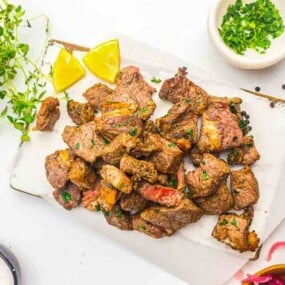 A serving of grilled steak bites garnished with herbs, accompanied by lemon wedges, a bowl of chopped herbs, and various seasonings on a white surface.