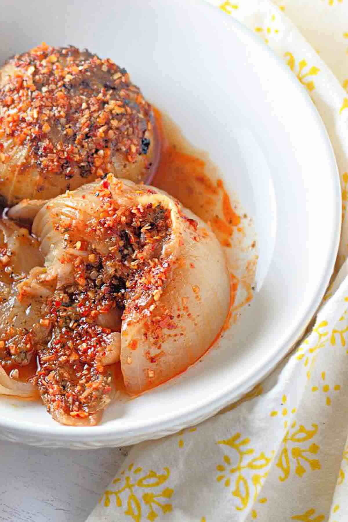 Two stuffed onions covered in a red sauce and spice mixture are served in a white bowl on a white and yellow patterned cloth.