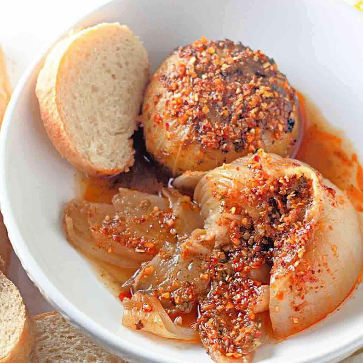A white bowl containing baked onions topped with chili flakes and spices, served with two slices of bread.